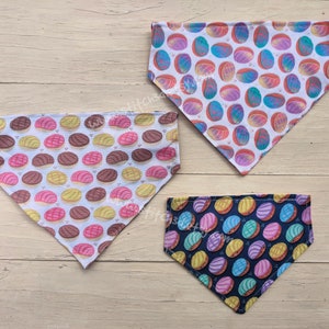 May include: Three dog bandanas with colorful concha bread patterns. One bandana has a white background with pink, brown, and yellow conchas. Another has a white background with blue, pink, and orange conchas. The third has a navy background with pink, yellow, and blue conchas.