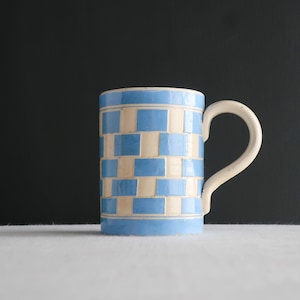 May include: A blue and white ceramic mug with a checkered pattern. The mug has a handle and is made in a rustic style.