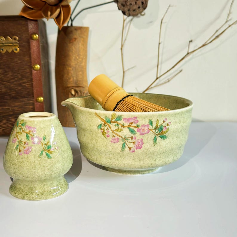 K&ouml;nnte beinhalten: Eine gr&uuml;ne Keramik-Matcha-Schale mit Blumenmuster und einem Bambus-Schneebesen. Die Schale hat einen Ausguss und eine passende gr&uuml;ne Keramik-Matcha-Tasse mit Blumenmuster.