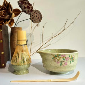 May include: A green ceramic matcha bowl with a floral design, a bamboo whisk with a green ceramic base, and a wooden matcha scoop.