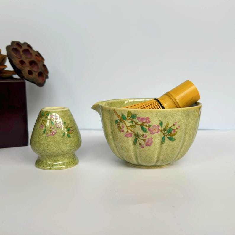 K&ouml;nnte beinhalten: Eine gr&uuml;ne Keramik-Matcha-Schale mit Blumenmuster und einem Bambus-Schneebesen. Die Schale hat einen Ausguss und eine strukturierte Oberfl&auml;che.