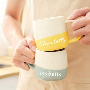 Könnte beinhalten: Zwei Keramikbecher, einer gelb und einer hellblau, aufeinander gestapelt. Auf dem oberen Becher steht der Name "Charlotte" in weißer Schrift, auf dem unteren "Isabella". Beide Becher haben eine cremefarbene obere Hälfte.