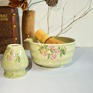 May include: A green ceramic matcha bowl with a floral design and a bamboo whisk. The bowl has a spout and a matching green ceramic matcha cup with a floral design.