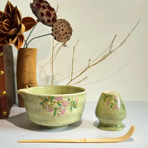 K&ouml;nnte beinhalten: Eine gr&uuml;ne Keramik-Matcha-Schale mit Blumenmuster, ein passender gr&uuml;ner Keramik-Schneebesenhalter und ein Bambus-Matcha-Schneebesen.