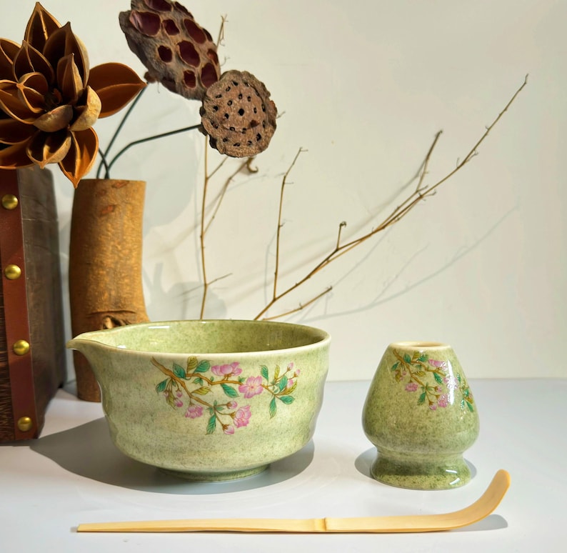 K&ouml;nnte beinhalten: Eine gr&uuml;ne Keramik-Matcha-Schale mit Blumenmuster, ein passender gr&uuml;ner Keramik-Schneebesenhalter und ein Bambus-Matcha-Schneebesen.