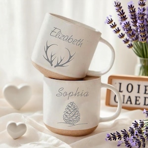 Könnte beinhalten: Zwei weiße Keramikbecher, die übereinander gestapelt sind. Der obere Becher trägt den Namen "Elizabeth" und ein Hirschgeweih-Design. Der untere Becher trägt den Namen "Sophia" und ein Tannenzapfen-Design. Beide Becher haben einen beigen Boden und einen weißen Henkel.