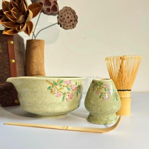 May include: A green ceramic matcha bowl with a floral design, a matching green ceramic matcha cup, a bamboo whisk, and a bamboo matcha scoop.