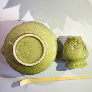May include: A green ceramic matcha bowl with a matching green ceramic whisk and a small green ceramic cup with a floral design.