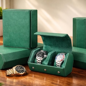 Puede incluir: Cajas y estuches de almacenamiento de relojes verde esmeralda. Se muestra un reloj dorado con esfera negra y un reloj plateado con esfera negra. Una caja de reloj verde está abierta, revelando dos relojes. También se ven otras cajas y estuches verdes.