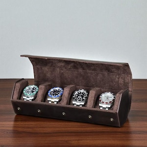 Peut inclure: Un étui à montres en cuir marron avec quatre compartiments contenant chacun une montre-bracelet argentée avec une lunette de couleur différente. L'étui est ouvert, révélant un intérieur marron doux. Les montres sont exposées.