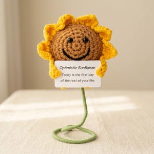 Può includere: Un girasole all'uncinetto con petali gialli e un centro marrone con un volto sorridente. Il girasole regge un cartoncino rettangolare bianco con il testo: Optimistic Sunflower. Today is the first day of the rest of your life. Stelo verde.