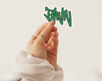 May include: A green sticker with the word "JAWN" in a graffiti-style font. The sticker is held up by a hand with rings on the fingers. The background is a plain white.