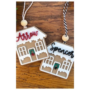 May include: Two gingerbread house ornaments with personalized names. One ornament says "Arrow" in red script, and the other says "Spencer" in black script. Each house has white frosting, windows, and a wreath. They hang from a string with a wooden bead.