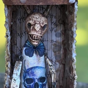 May include: A handcrafted, spooky art piece featuring a skeletal figure inside a rustic box. The figure has a skull-like head, a blue bow tie, and a blue skull graphic on its torso. The box is labeled "DO NOT OPEN".