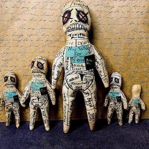 May include: A collection of five handmade voodoo dolls. The dolls are beige with black and red stitched details, including facial features. Each doll has a turquoise rectangle with the words "My Wish For You". The dolls are labeled with various body parts and ailments.