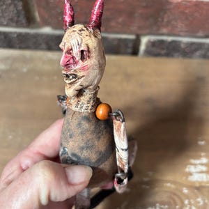 May include: A handmade doll with a devilish appearance. The doll has a pale face with red horns, a stitched mouth, and a printed fabric collar. The body is made of fabric and the arms are articulated. An orange bead is attached to the neck.