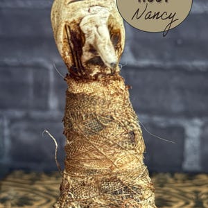 May include: A handcrafted, unique doll with a weathered, aged appearance. The doll's head is off-white with a prominent nose and mouth. The body is wrapped in textured, tan fabric. The text "NOSY Nancy" is in a brown oval.