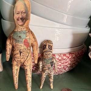 May include: Two handmade fabric dolls. The larger doll has a photo of a woman's face on the head and text on the body, including "My Wish For You" and "Broken Heart." The smaller doll has a skull face and similar text. The dolls are tan with black text.
