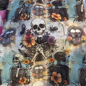 May include: A fabric with a repeating pattern of skeletons, flowers, butterflies, and spiders. The skeletons are depicted in various poses, surrounded by orange, purple, and pink flowers. The background is a light blue with a distressed, vintage look.