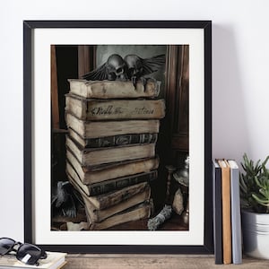 May include: A framed print showing a stack of old books, with the words "Skull & Initiation" on one. Two skull sculptures with wings are on top. The artwork has a gothic or occult theme, with a dark, vintage aesthetic.