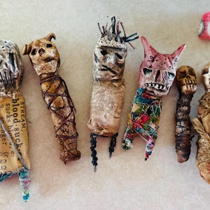 Peut inclure: Une collection de six figurines faites à la main, fantaisistes, avec des visages en forme de crâne. Les figurines sont fabriquées à partir de divers matériaux, dont du tissu, de la ficelle et du papier. Une figurine porte le texte "blood sucks". Les figurines sont de couleurs variées.