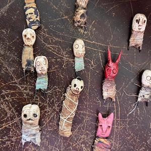 Peut inclure: Une collection de petites figurines faites à la main, ressemblant à des poupées, avec des visages sculptés et des corps enveloppés. Certaines sont peintes en rouge ou rose, d'autres sont en bois naturel. Les figurines ont diverses expressions et sont disposées sur une surface sombre et texturée.