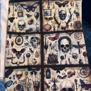 May include: A fabric panel featuring a repeating pattern of skulls, butterflies, insects, and antique-style illustrations. The design incorporates a vintage aesthetic with a muted color palette of beige, black, and blue. The panel is divided into rectangular sections.