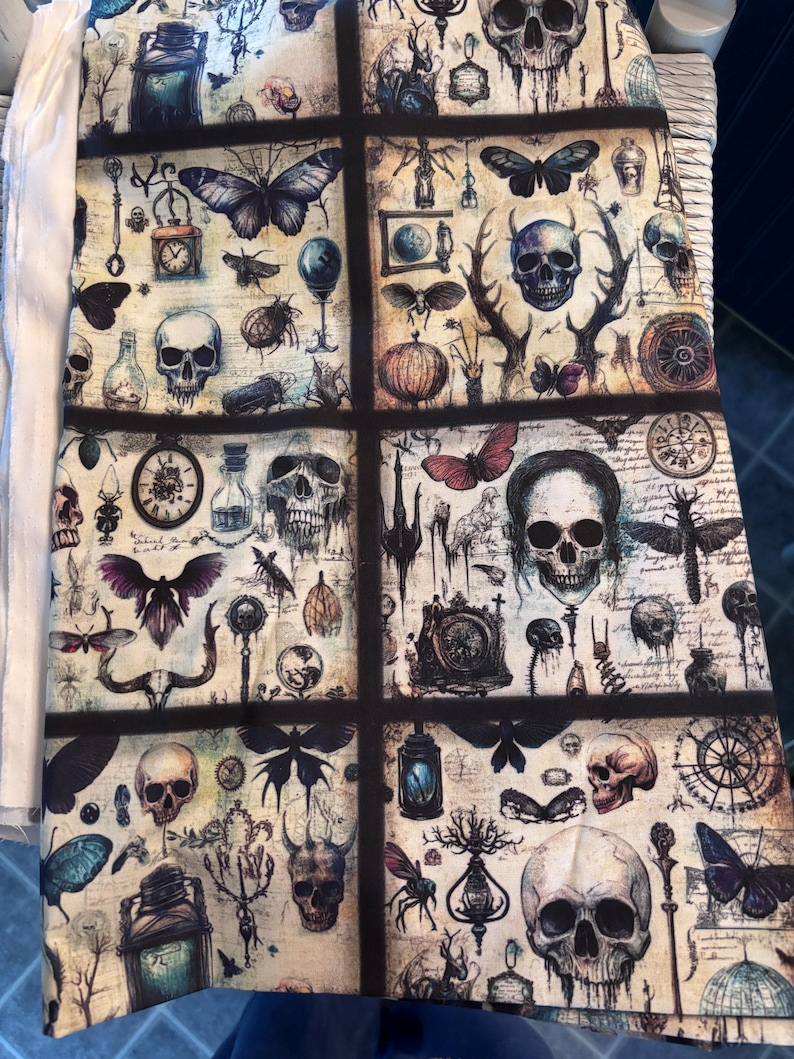 May include: A fabric panel featuring a repeating pattern of skulls, butterflies, insects, and antique-style illustrations. The design incorporates a vintage aesthetic with a muted color palette of beige, black, and blue. The panel is divided into rectangular sections.
