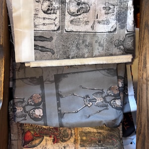 May include: A collection of fabric pieces with anatomical illustrations of skeletons and organs. The fabric features a grayscale color palette with detailed drawings of skulls, bones, and internal organs. The fabric is stacked inside a wooden drawer.