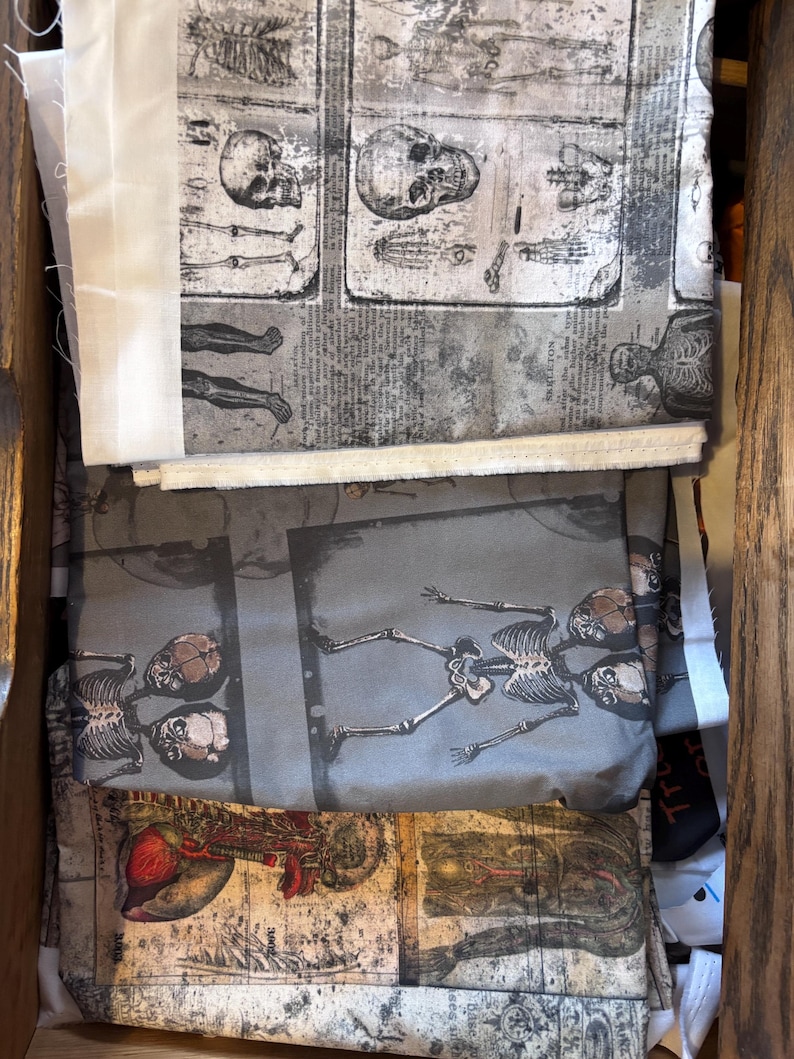 May include: A collection of fabric pieces with anatomical illustrations of skeletons and organs. The fabric features a grayscale color palette with detailed drawings of skulls, bones, and internal organs. The fabric is stacked inside a wooden drawer.