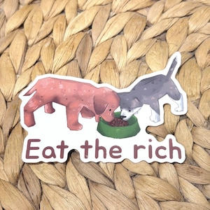 May include: A die-cut sticker featuring a pink dog and a gray dog eating from a single green bowl. The text "Eat the rich" is printed in brown letters below the dogs. The illustration has a white border and a matte finish on the surface.