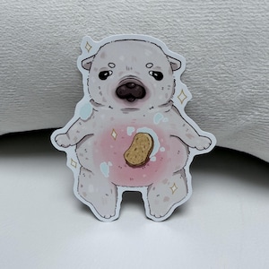 Könnte beinhalten: Ein gestanzter Aufkleber mit einem Cartoon-Wombat. Das Wombat hat einen hellgrauen Körper, einen rosa Bauch und einen braunen Gegenstand in der Mitte. Es hat kleine schwarze Augen und ist von blauen Blasen und funkelnden Akzenten umgeben.