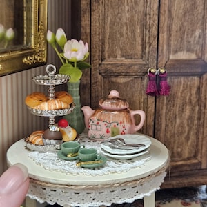 May include: Miniature tea party scene with a small round table set with a lace tablecloth, two green teacups, plates, and a teapot. A tiered serving tray holds pastries. A vase with flowers and a wooden cabinet are in the background.