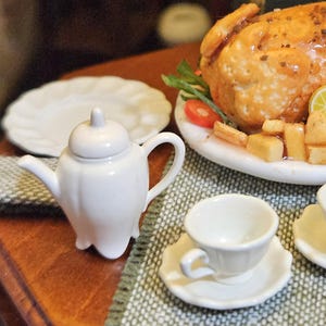 May include: A miniature white ceramic teapot, two small teacups on matching saucers, and a white plate with a scalloped edge. Beside these items sits a larger plate featuring a roasted chicken, sliced lemon, and diced potatoes.