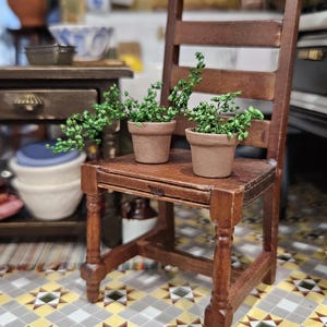 May include: A miniature wooden chair with a dark brown finish, holding two small terracotta pots with green plants. The chair is placed on a patterned tile floor, with other miniature furniture in the background.