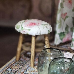 May include: A small, round stool with a floral rose pattern on the seat. The seat is white with pink and green roses. The legs are made of light brown wood. Clear glass jars are in the foreground.