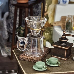 May include: Miniature glass coffee maker with a gold-colored metal filter, a small glass carafe, and two green teacups with saucers. A wooden coffee grinder and a wooden cutting board are also visible. The scene is set on a miniature wooden table.