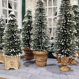 May include: Miniature Christmas trees with snow-like accents in decorative pots. The trees are green with white tips, and the pots are beige with ornate designs. The scene is set on a gray brick-like surface, evoking a festive holiday theme.