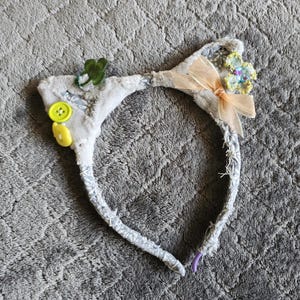 May include: A gray cat ear headband with a floral and bow embellishment. The headband features a light gray fabric with a floral pattern, yellow buttons, and a green shamrock. The ears are decorated with a peach-colored bow and a cluster of small flowers.