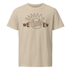 Puede incluir: Una camiseta beige con un dibujo de línea marrón de una cara con una corona de púas y el texto "WE EN".
