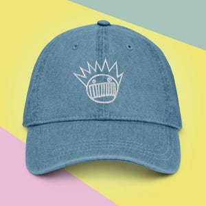 Puede incluir: Una gorra de béisbol de mezclilla azul con una imagen bordada en blanco de una cara de dibujos animados con una corona de púas.