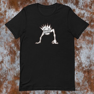 Op de afbeelding: Zwart T-shirt met een witte afbeelding van een cartoonfiguur met een mond en stekelig haar. Het figuur heeft lange armen en benen.
