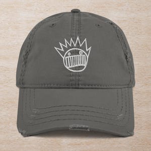 May include: A grey distressed baseball cap with a white embroidered image of a cartoon face with a crown of spikes.