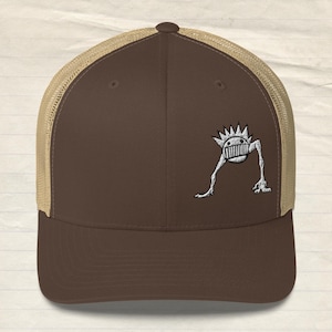 Puede incluir: Gorra de camionero marrón y beige con un gráfico blanco bordado de una criatura de dibujos animados con una cara y cuatro patas.