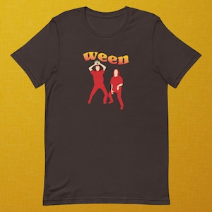 Könnte beinhalten: Dunkelbraunes T-Shirt mit dem Wort "ween" in orange-gelber Retro-Schrift. Darunter befinden sich zwei rote Figuren in Tanzpose. Das Shirt besteht aus einem weichen Material.