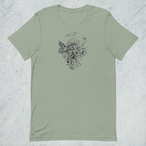May include: A sage green t-shirt with a black ink illustration of a surreal design. The artwork features feathers, a claw, and other abstract shapes. The shirt has a classic crew neck and short sleeves. The fabric appears soft.