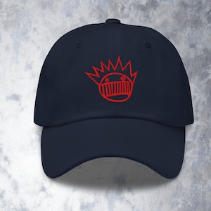 May include: Navy blue baseball cap with a red embroidered design of a face with a crown. The cap has a curved brim and an adjustable strap at the back. The design is a simple, cartoon-like illustration.