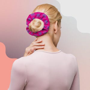 May include: A vibrant pink scrunchie, patterned with blue motifs, encircles a bun hairstyle. The scrunchie, made from soft material, adds a splash of colour to the hair. The background features a gradient of pink and white hues.