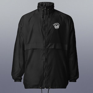 May include: A black windbreaker jacket with a white graphic of a cartoon face with a crown on top. The jacket has a zipper closure and two pockets.