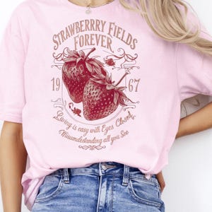 May include: A light pink t-shirt featuring a maroon graphic with the words "Strawberry Fields Forever" and two strawberries. The graphic also includes the numbers "1967" and the phrase "Living is easy with eyes closed, misunderstanding all you see."